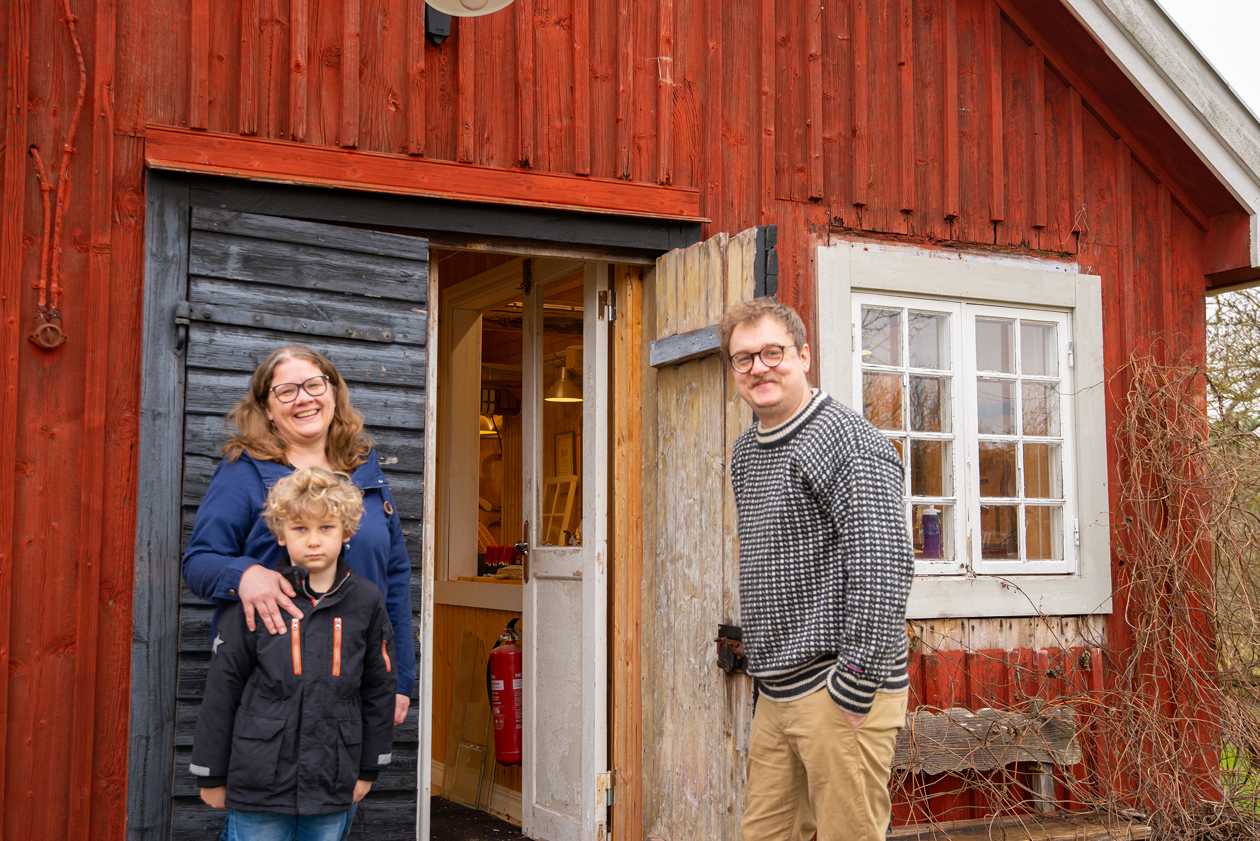 Lars och Åsa samt deras son står på trappan och håller upp dörren till verkstaden. 